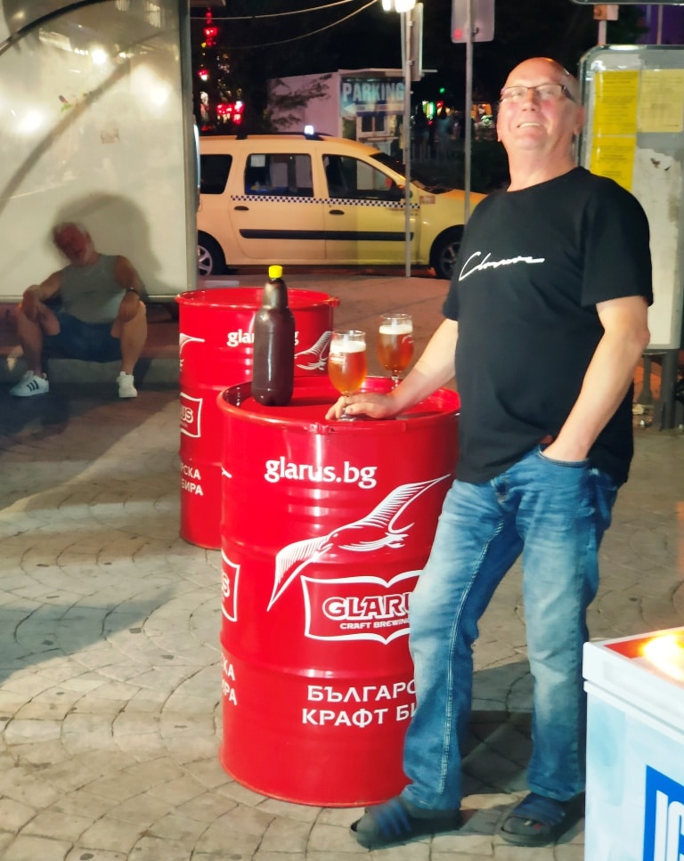 Mężczyzna zadowolony z butelką piwa kraftowego Glarus opiera się o beczkę z logo browaru